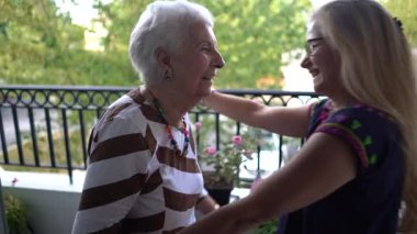 Gülerken, yaşlı bir kadın ve genç bir kadın balkonda birbirlerine sarılıyorlar..