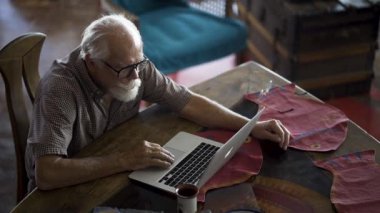 Beyaz sakallı, yemek masasında dizüstü bilgisayarda çalışan ve yanında kahve fincanı olan yaşlı bir adama tepeden bakıyorum..