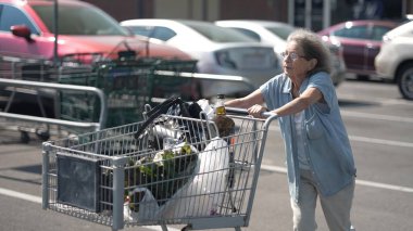 Yaşlı, yaşlı bir kadın haftalık alışverişinden sonra bakkal arabasıyla otoparkta geziniyor..