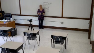 Geniş açılı, gülümseyen İngilizce öğretmeni, boş sandalyeli bir sınıfın beyaz tahtasına yazı yazıyor..