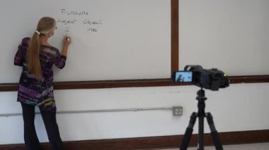 Bir okul sınıfında beyaz tahtaya yazı yazan kadın İngilizce öğretmeni video kameranın önünde uzaktan melez öğrenimi için tıbbi bir maske takıyor..