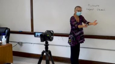 Bir okul sınıfında beyaz tahtaya yazı yazan kadın İngilizce öğretmeni video kameranın önünde uzaktan melez öğrenimi için tıbbi bir maske takıyor..