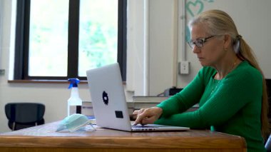 Okulda öğretmenlerin yan görüntüsü. Masasında tıbbi yüz maskesi ve masasında PPE ve ABD bayrağı olan bilgisayar üzerinde çalışıyorlar..