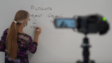 Bir okul sınıfında beyaz tahtaya yazı yazan kadın İngilizce öğretmeni video kameranın önünde uzaktan melez öğrenimi için tıbbi bir maske takıyor..