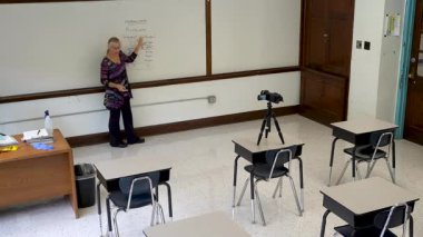 Geniş açılı, gülümseyen İngilizce öğretmeni, boş sandalyeli bir sınıfın beyaz tahtasına yazı yazıyor..