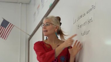 Güzel sarışın bir kadın, beyaz tahtada İspanyolca öğretiyor. Üzerinde Amerikan bayrağı asılı..