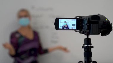 Bir okul sınıfında beyaz tahtaya yazı yazan kadın İngilizce dil bilgisi öğretmeninden video kameranın önünde uzaktan melez öğrenimi için tıbbi bir maske takmaya odaklan..