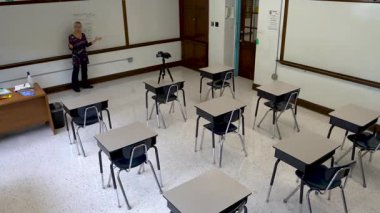 Geniş açılı, gülümseyen İngilizce öğretmeni, boş sandalyeli bir sınıfın beyaz tahtasına yazı yazıyor..