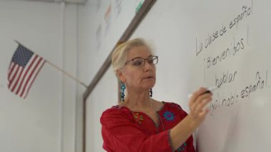 Güzel sarışın bir kadın, beyaz tahtada İspanyolca öğretiyor. Üzerinde Amerikan bayrağı asılı..
