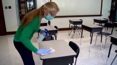 Ağır çekimde, tıbbi yüz maskesi takan bir bayan öğretmen, covid-19 sırasında boş bir sınıfta sandalye ve masayı dezenfekte eder..