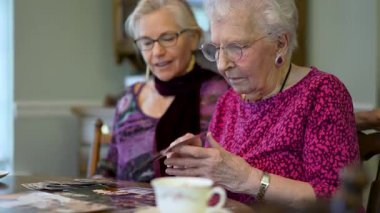 Yaşlı, gülümseyen bir kadın eski fotoğraflara bakıyor ve yemek masasında kızıyla olan anılarını hatırlıyor..