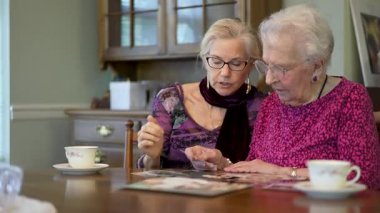 Kamera, yaşlı, gülümseyen bir kadının eski fotoğraflara bakmasını ve yemek masasındaki kızıyla ilgili anıları hatırlamasını sağlıyor..