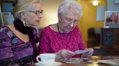 Yaşlı ve gülümseyen bir kadının eski fotoğraflara bakıp yemek masasında kızıyla olan anılarını hatırladığı yakın plan..