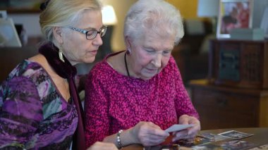 Yaşlı, gülümseyen bir kadının eski fotoğraflara bakması ve kızıyla yemek masasında geçirdiği anıları hatırlaması..