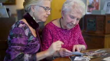 Yaşlı ve gülümseyen bir kadının eski fotoğraflara bakıp yemek masasında kızıyla olan anılarını hatırladığı yakın plan..