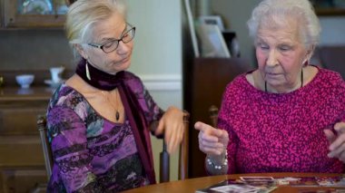 Yaşlı, gülümseyen bir kadın eski fotoğraflara bakıyor ve yemek masasında kızıyla olan anılarını hatırlıyor..