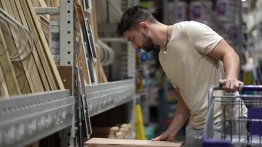 Sakallı genç bir adam hırdavatçıda ya da depoda kereste seçiyor..