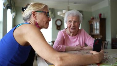 Mutlu gülümseyen, gülen yaşlı bir kadın olgun orta yaşlı bir kızla masada oturuyor, telefonda görüntülü sohbet ediyor. Gülümseyen sarışın bayan akıllı telefondan aileyi dinliyor..