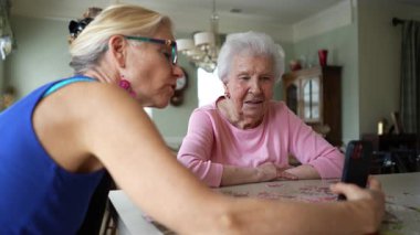 Mutlu gülümseyen, gülen yaşlı bir kadın olgun orta yaşlı bir kızla masada oturuyor, telefonda görüntülü sohbet ediyor. Gülümseyen sarışın bayan akıllı telefondan aileyi dinliyor..
