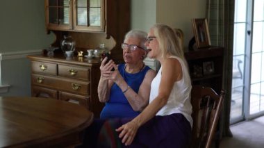 Yemek odasında yaşlı bir kadın ve yetişkin bir kız gözlüklü ve akıllı telefonlara bakarak eğleniyorlar..