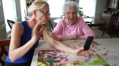 Mutlu gülümseyen, gülen yaşlı bir kadın olgun orta yaşlı bir kızla masada oturuyor, telefonda görüntülü sohbet ediyor. Gülümseyen sarışın bayan akıllı telefondan aileyi dinliyor..