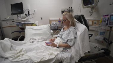 Winchester, VA, ABD - 09 03 2025: Bir hastane acil servisinde, yetişkin bir kadın hastane önlüğü giymiş bir şekilde yatakta oturuyor. Tedavi görürken telefonuna odaklanmış..