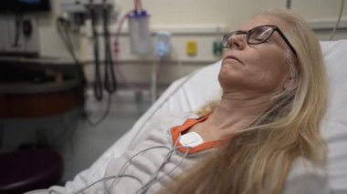 A mature woman is resting in a hospital room while connected to heart and EKG monitors. She appears calm and receives medical attention during her stay.