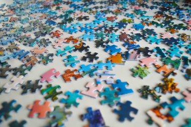 Yapboz parçalarının yüksek çözünürlüklü görüntüsü beyaz bir masaya saçılmış. Kaotik düzenleme zorlukları, düzensizliği ve çözüm bulma sürecini sembolize eder. Bu resim yaratıcılığı, problem çözmeyi, organizasyonu ve