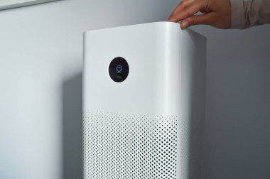 Minimalist bir iç mekanda gösterişli, modern bir hava temizleyicisi, üzerine nazikçe yerleştirilmiş bir el. Dijital ekranda, temiz, temiz havanın evdeki önemini vurgulayan hava kalitesi seviyeleri görülüyor. Bu cihaz sağlıklı bir yaşam tarzını, kapalı alanda geliştirilmeyi simgeliyor.