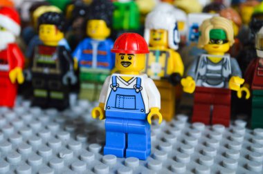 Tek bir LEGO çalışanı minyatür figür önplanda güven içinde duruyor, liderlik, bireysellik ve kararlılığı sembolize ediyor. Arkalarında geniş, çeşitli LEGO minyatürlerinden oluşan bir kalabalık takım çalışması, toplum ve ortak çabayı temsil ediyor. Bu yaratıcı