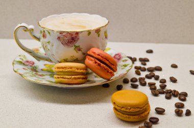 Köpüklü kahveyle dolu, renkli badem kurabiyelerinin yanında çiçek tabağında servis edilen hoş bir çay fincanı. Dağılmış kahve çekirdekleri sahneyi tamamlayarak sıcak ve davetkar bir atmosfer yaratıyor. Kahve ve tatlı aşkı için mükemmel bir an.