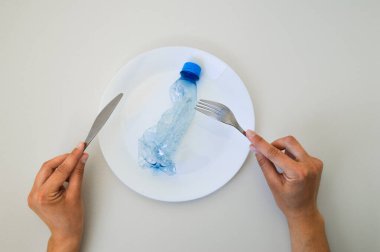 Kavramsal bir görüntü, temiz beyaz bir masa üzerinde çatal ve bıçak arasına yerleştirilmiş, buruşmuş plastik bir şişeyle beyaz bir tabak gösteriyor. Çatal bıçak takımını tutan eller tipik bir yemek takımını işaret ediyor ama plastiğin varlığı endişe verici gerçeği vurguluyor.