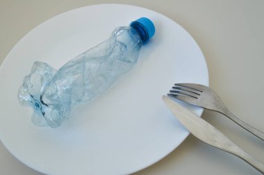 Tek bir çatalla beyaz bir tabakta servis edilen buruşmuş plastik şişeyi yemek olarak gösteren güçlü bir görsel kavram. Bu minimalist ama çarpıcı görüntü modern insan diyetinde mikroplastiğin varlığını sembolize eder. Bu dikkat çeker.