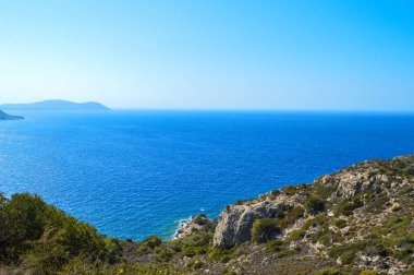 Yunanistan 'ın Rodos kıyı şeridi boyunca uzanan derin mavi Ege Denizi' nin nefes kesici manzarası. Sahne engebeli kayalıklar ve vahşi Akdeniz bitkileri tarafından çerçevelenmiş, gözü denizin gökyüzü ile buluştuğu sakin ufka götürüyor. Mesafe