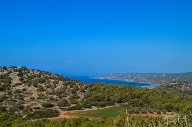 Yunanistan 'ın Rodos kıyı şeridi boyunca uzanan derin mavi Ege Denizi' nin nefes kesici manzarası. Sahne engebeli kayalıklar ve vahşi Akdeniz bitkileri tarafından çerçevelenmiş, gözü denizin gökyüzü ile buluştuğu sakin ufka götürüyor. Mesafe