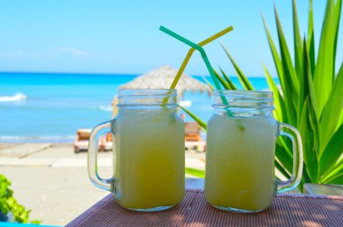 Mason kavanozlarındaki iki ferahlatıcı limonata plaj kenarındaki masada mükemmel bir şekilde soğutulmuş, pipetleri neşeyle güneşin altında birbirine dolanmış. Ege Denizi 'nin berrak turkuaz suları ve arka planda palmiye püsküllü bir şemsiye ile bu fotoğraf...