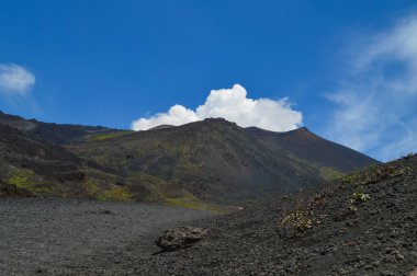 İtalya 'nın Sicilya kentindeki Etna Dağı' nın volkanik yamaçlarının manzarası. Siyah lav kayaları, seyrek bitki örtüsü ve canlı mavi gökyüzü patlamalarla şekillenen dramatik bir doğal manzara yaratır..