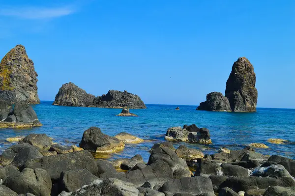 İtalya 'nın Sicilya kıyısındaki Aci Trezza yakınlarındaki mavi sulardan yükselen volkanik deniz yığınları. Açık gökyüzü ve engebeli kaya oluşumları sakin bir Akdeniz manzarası yaratır.