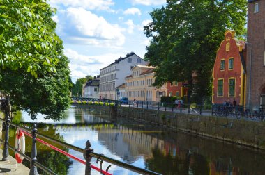 Bereketli yeşil ağaçlar ve uzak bir köprüyle çerçevelenmiş sakin sulara yansıyan tarihi binalarla dolu Stockholm kanalının büyüleyici bir manzarası. Güneşli bir günde şehrin dingin güzelliğini yakalar. Seyahat ve Avrupa şehir manzaraları için ideal