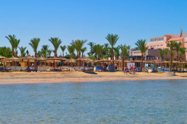 Palmiye ağaçları, geleneksel plaj şemsiyeleri ve arka planda kristal berrak turkuaz su ve otelle altın kumda dinlenen tatilcilerin yer aldığı güzel bir tropikal plaj beldesi.