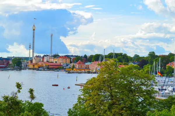 Stockholm 'ün büyüleyici limanının manzarası canlı renkli binalar, yelkenli marinalar ve dramatik bulutlu gökyüzü altında yemyeşil yeşillikler. İskandinav seyahat içeriği için mükemmel