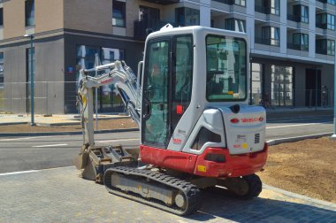 Temiz kırmızı ve beyaz bir Takeuchi mini kazıcısı, arka planda modern apartmanlar bulunan kentsel bir inşaat sahasının asfaltlı bir yüzeyinde hazır bekliyor. Bu resim verimliliği, inşaat endüstrisini ve kentsel gelişimi vurguluyor