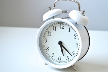 Temiz bir yüzeye zamansız beyaz analog alarm saatinin çıtır çıtır bir yakın çekimi. Bu görüntü zaman kavramlarını, dakikliği, uyandırma çağrılarını, teslim tarihlerini ve basit, klasik bir estetiği çağrıştırıyor..