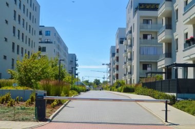 Otomatik patlama bariyeri, çağdaş apartman binaları ve yeşil peyzaj ile çevrili Varşova 'daki modern bir yerleşim alanına erişimi kontrol ediyor. Olay yeri, güvenlik ve denetimli şehir yaşamını vurguluyor