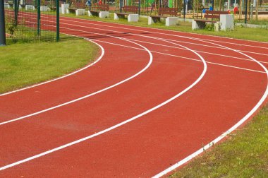 Yeşil spor alanında kıvrımlı beyaz şeritli canlı bir kırmızı koşu pisti, arka planda banklar var. Bu resim fitness, atletizm, açık hava eğlencesi ve şehir spor tesislerini sembolize eder.