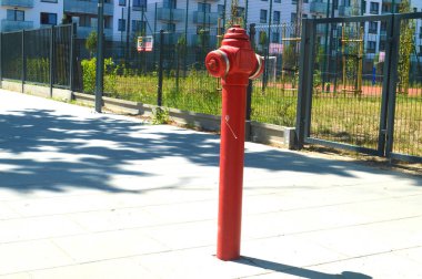 Parlak kırmızı bir yangın musluğu, muhtemelen Varşova 'nın kentsel bir ortamında çitlerle ve modern konut binalarıyla çevrili asfalt bir geçitte belirgin bir şekilde duruyor. Şehir altyapısını ve acil servisleri sembolize eder.