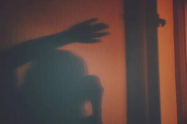Korku sahnesinde cam kapının arkasındaki kadın, korkunç cadılar bayramı konsepti. Duvardaki gölge. Balkondaki kız. Gün batımı. Turuncu ışık. Yatay fotoğraf.