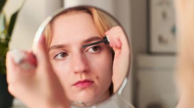 Kadın büyüteçle bakıyor. Genç, açık tenli bir kız kaşlarını boyuyor. Geniş kaşlar. Doğal güzellik. Mükemmel hafif makyaj. İnce pembe dudaklar. Aynadaki yansıma. Kaşlar için jel..