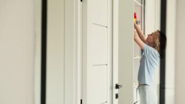 Beyaz elbiseli genç kadın dolapta duruyor. Sarışın kız dolaptan bir kutu çıkartıyor. Üst rafta. Koridordaki aynadaki yansıma.