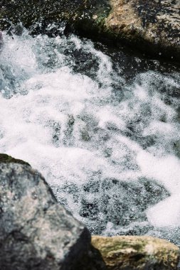 Ormanda şelale. Deniz. Su sıçrıyor. Kıyıya. Dalgalar. Deniz köpüğü. Denizin ortasında taş var. Kum. Deniz yosunu. Yaz tatili. Deniz kıyısında bir tatil köyü. Dalgalar. Deniz yosunlu taşlar.
